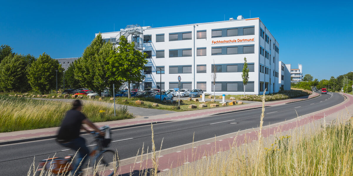 Building photo with "Fachhochschule Dortmund" logo on building 38b - from a distance. Cyclist enters the picture on the left.