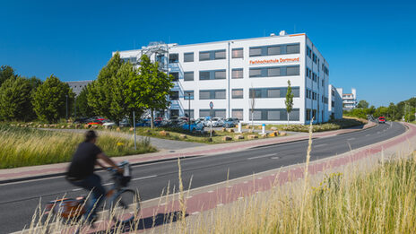 Building photo with "Fachhochschule Dortmund" logo on building 38b - from a distance. Cyclist enters the picture on the left.