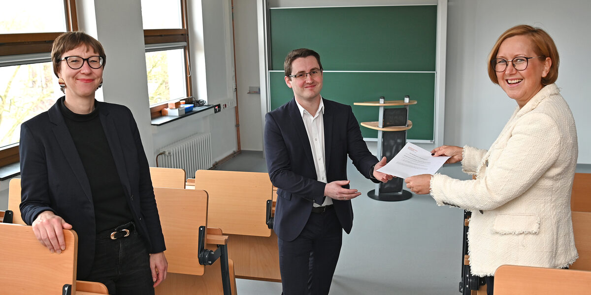 Drei Personen stehen in einem Hörsaal. Einer Person bekommt ein Stück Papier überreicht.