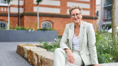Eine Frau in einem grünen Blazer sitzt draußen vor einem Gebäude und lacht.__A woman in a green blazer sits outside in front of a building and laughs