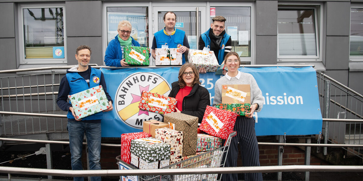 Sechs Personen stehen auf dem Aufgang zum Eingang der Bahnhofsmission Dortmund. Jede von ihnen hält ein festlich verpacktes Päckchen in den Händen. Vorn in der Mitte steht ein Einkaufswagen voll mit weiteren Geschenkpäckchen.
