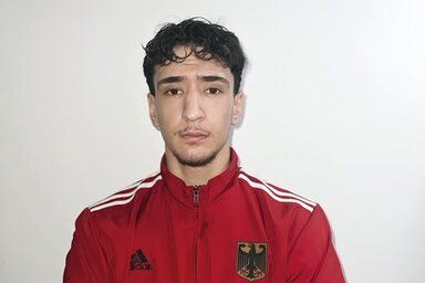 Ein Mensch in roter Trainingsjacke mit dem deutschen Wappen auf der Brust schaut mit verschränkten Armen in die Kamera.