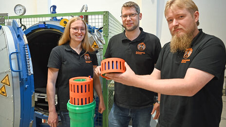 Drei Personen stehen im Hochspannungslabor der FH Dortmund und zeigen ein Bauteil eines Batteriespeichers.