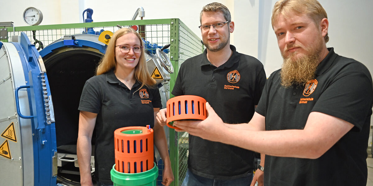 Drei Personen stehen im Hochspannungslabor der FH Dortmund und zeigen ein Bauteil eines Batteriespeichers.