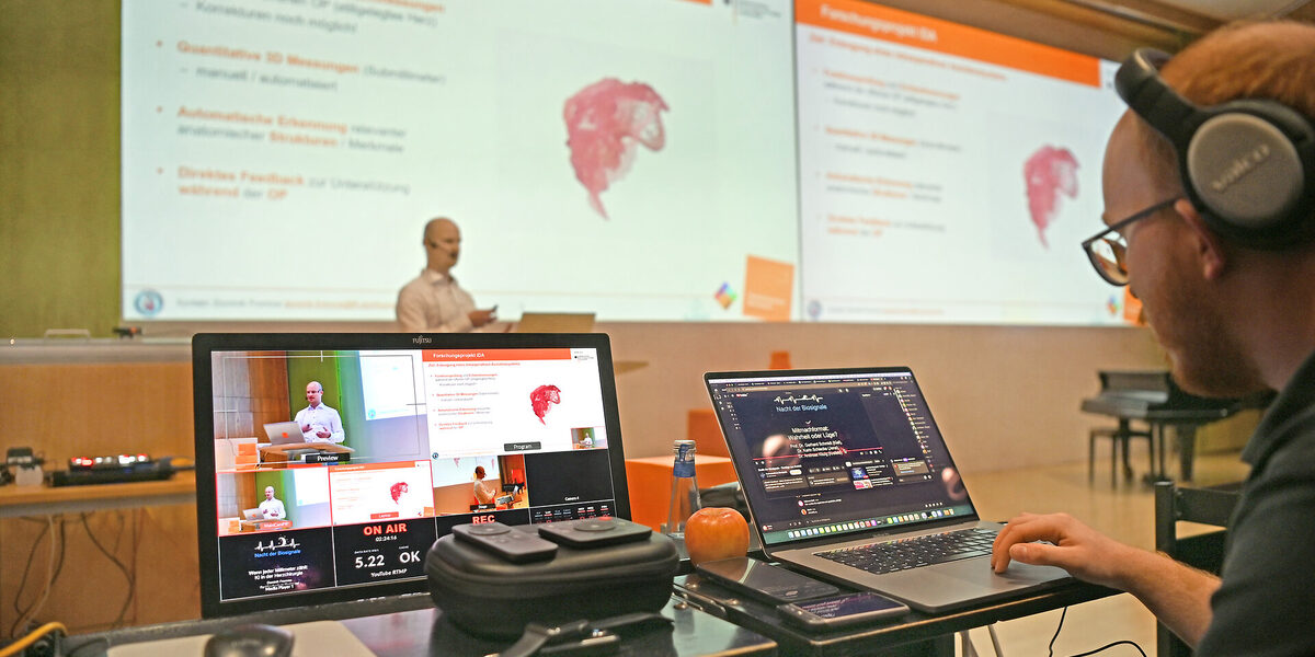 Eine Person hält einen Vortrag, im Vordergrund sind Monitore zu sehen. Der Vortrag wird live ins Netz gestreamt.