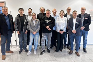 Elf Personen stehen nebeneinander vor einer hellen Wand und schauen freundlich in die Kamera.