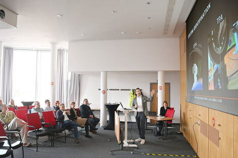 Eine Person steht am Rednerpult und weist auf die Projektion an der Wand rechts, links sitzen mehrere Personen und hören zu.