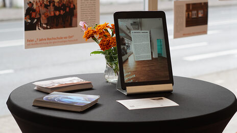 Auf einem Stehtisch befindet sich neben Flyern ein aufgestelltes iPad und eine Vase mit orangenen Blumen