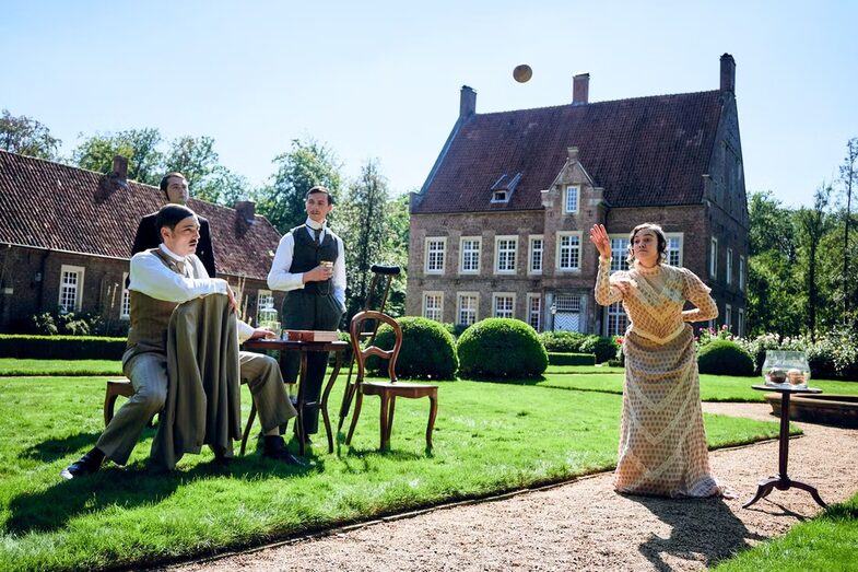 Eine Frau wirft einen Ball vor einem historischen Gebäude. Drei Männer beobachten sie kritisch.