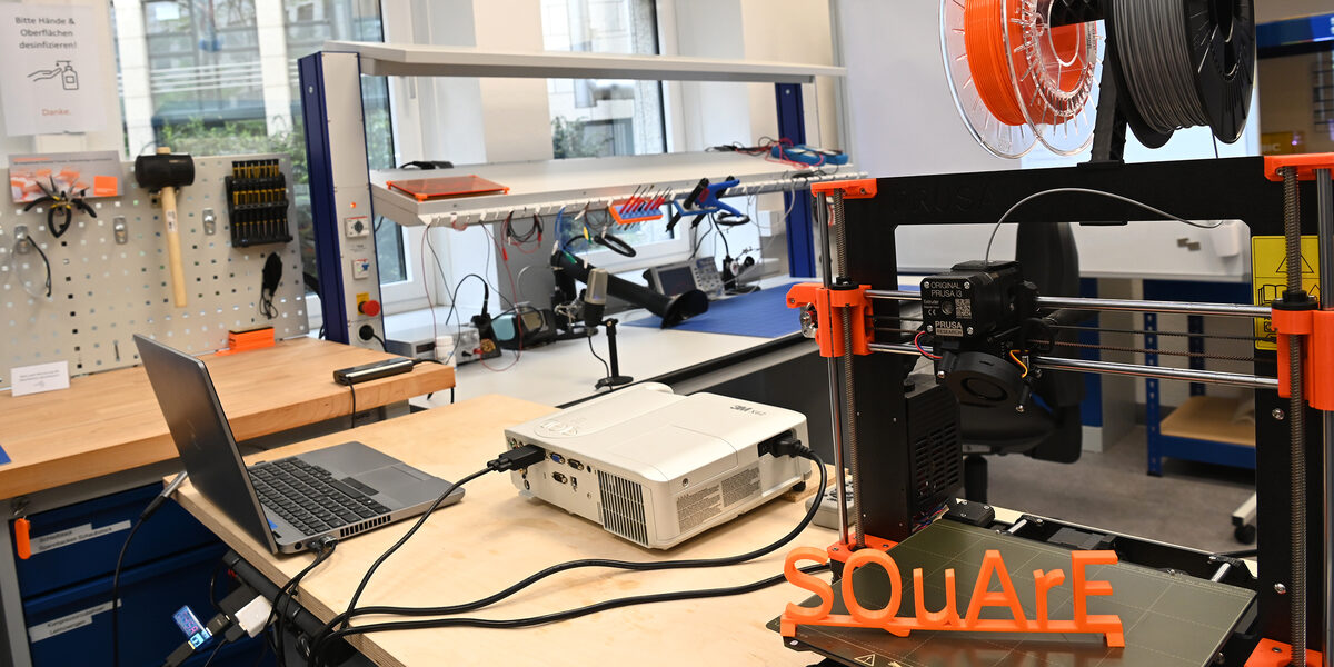 The picture shows a 3D printer with the printed three-dimensional SQuArE train; a workbench can be seen in the background.