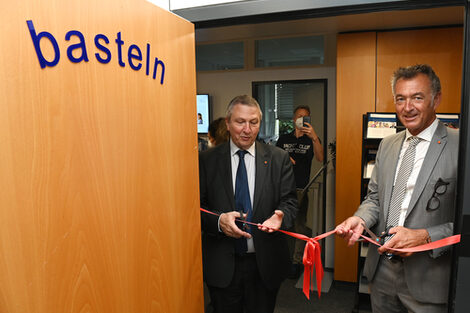 Two male figures cut a red ribbon hanging in a door frame. The door bears the inscription "tinker".