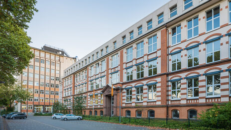 Foto des Altbaus und des Hochhauses an der Sonnenstraße mit Parkplatz davor. __ <br>View of the old building and the skyscraper on Sonnenstrasse across the parking lot.