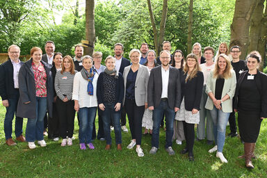 23 Personen stehen auf einer Wiese unter Bäumen und vor Büschen, durch die Baumkronen fällt Sonnenlicht.