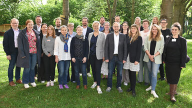 23 Personen stehen auf einer Wiese unter Bäumen und vor Büschen, durch die Baumkronen fällt Sonnenlicht.