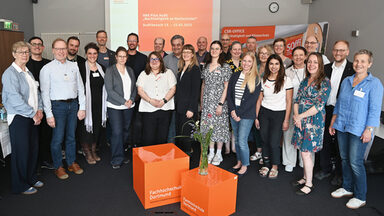 26 people stand in a semi-circle in a room in front of a presentation entitled "HRK pilot Aufdit "Sustainability at universities".