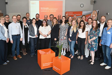 26 people stand in a semi-circle in a room in front of a presentation entitled "HRK pilot Aufdit "Sustainability at universities".