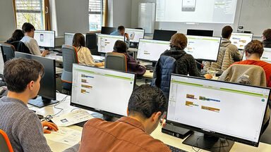 Mehrere Personen sitzen in einem Raum, vor ihnen stehen Computerbildschirme auf den Tischen.