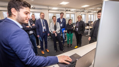 A male read person shows something on a laptop monitor to a larger group of people