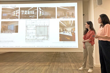 Two female characters stand in front of a screen and present an architectural design.