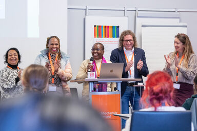 Fünf Personen stehen an einem Redenpult, die mittlere hält ein Mikro, die anderen klatschen, alle lächeln. Im Vordergrund sind die Hinterköpfe sitzender Menschen zu sehen.