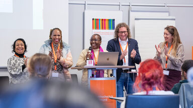 Five people are standing at a lectern, the middle one is holding a microphone, the others are clapping, everyone is smiling. The backs of the heads of seated people can be seen in the foreground.