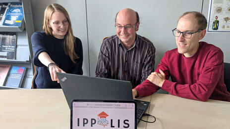 Three people are sitting in front of a laptop. The "IPPOLIS" logo can be seen on a monitor in front of them.