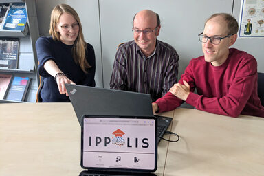 Drei Personen sitzen vor einem Laptop. Auf einem Monitor davor ist das Logo „IPPOLIS“ zu sehen.