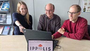 Drei Personen sitzen vor einem Laptop. Auf einem Monitor davor ist das Logo „IPPOLIS“ zu sehen.