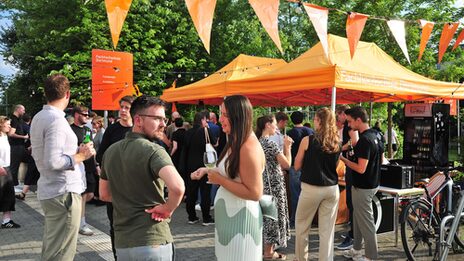 Menschen stehen auf dem Sommerfest des Fachbereichs Architektur der FH Dortmund. Unter orangefarbenen Pavillons versorgt die Fachschaft die Gäste mit Getränken.