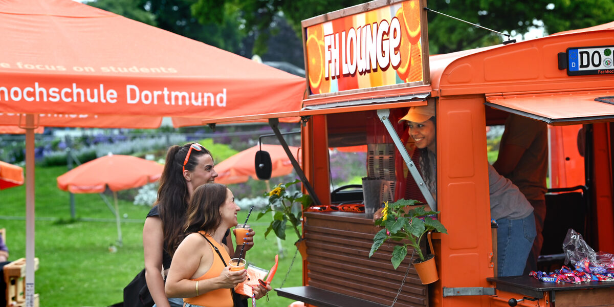 A sign reading "FH Lounge" is attached to the roof of an orange vintage car. From the interior, a person looks outside and smiles at two people standing in front of an open bar in the car, each holding a cup of slush ice, among other things. A meadow with orange parasols can be seen in the background.