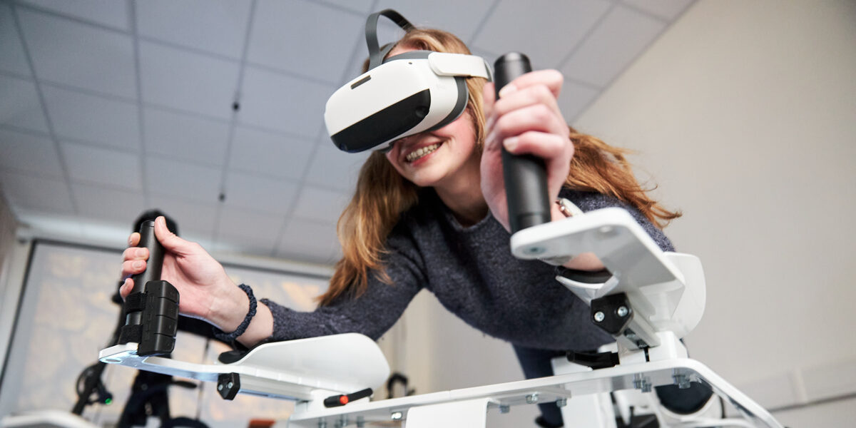 Foto von einer Person auf dem Icaros-Gerät. Sie trägt eine VR-Brille und hält sich an den Griffen fest.