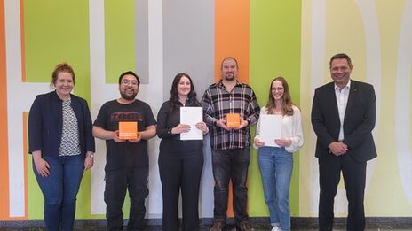 Six people stand in a row in front of a wall with brightly colored areas. Two people are holding a FH cube in front of them, two people are holding a white folder.