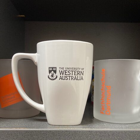 Zwei Tassen mit FH Dortmund in einem Regal mit einer Tasse mit dem Logo der UWA