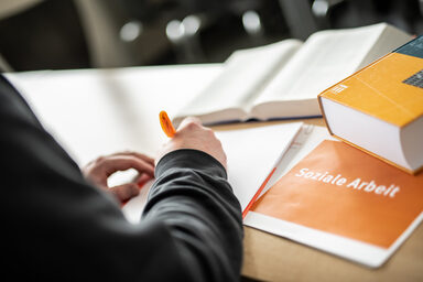 Cropped photo of a person writing something on a pad with an orange pen. Next to it are books and a notebook labeled "social work".