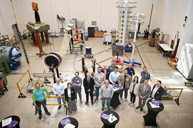 16 people, photographed from an elevated position, stand in a high hall surrounded by technical equipment and a few black-clad bar tables with brochures on them.