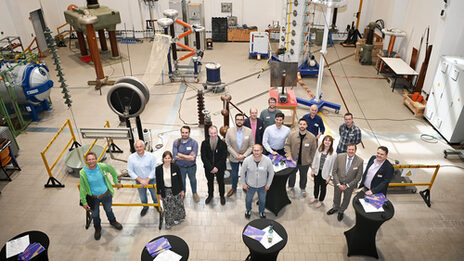 16 people, photographed from an elevated position, stand in a high hall surrounded by technical equipment and a few black-clad bar tables with brochures on them.