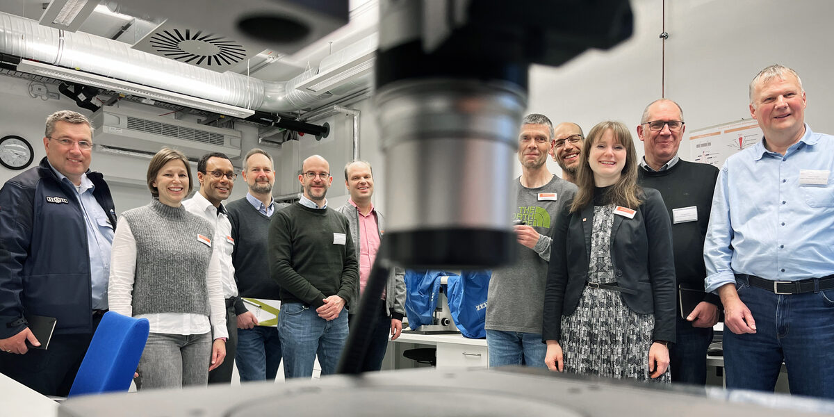Eleven people stand in a room facing the camera. In the center foreground, a silver cylinder protrudes into the picture from above, very close and out of focus. In the lower foreground, just as close, a dark plate can be seen.