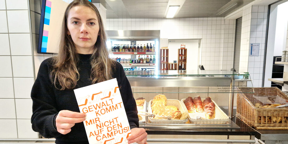 Eine Person steht in der Mensa vor einer Theke mit Backwaren und hält eine Tüte vor sich mit der Aufschrift: "Gewalt kommt mir nicht auf den Campus!".