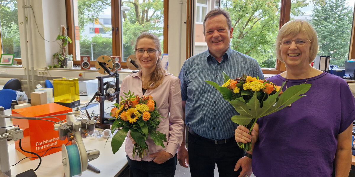 Three people stand next to each other in a room, two of them each holding a bouquet of flowers. Behind them, several objects are placed on tables, including 3D printers and a UAS cube.