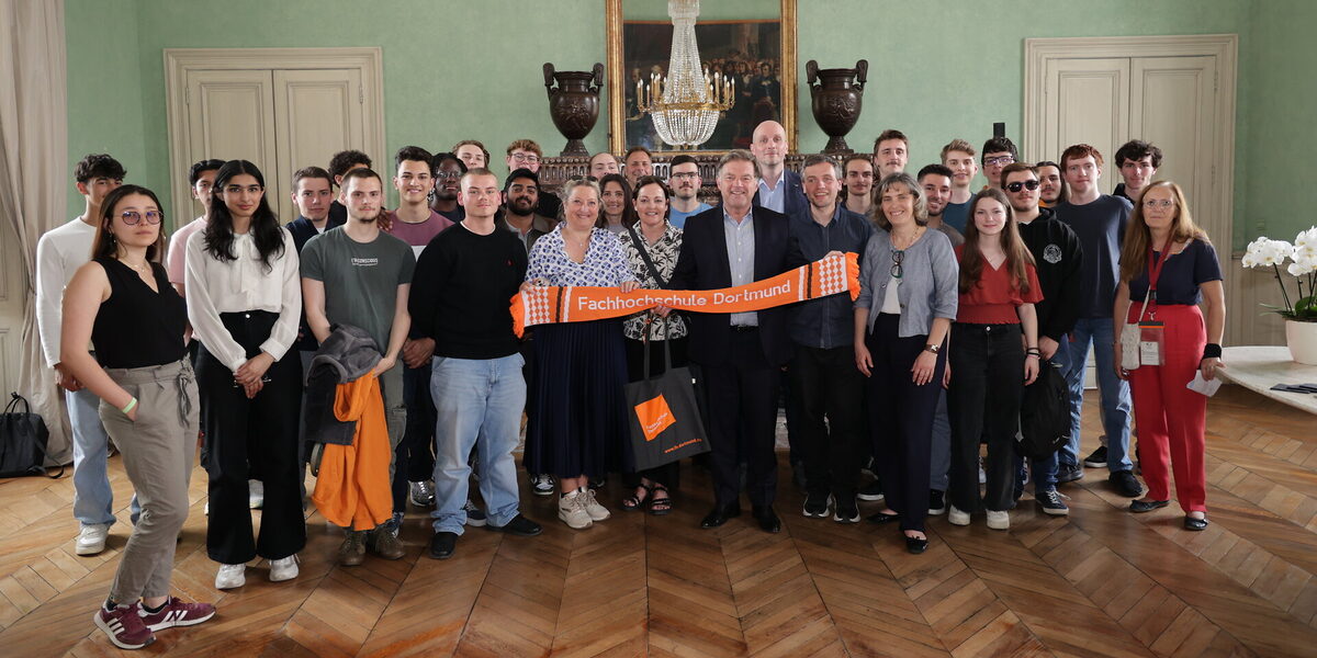 Eine Gruppe von Personen steht in einem Saal, die Personen in der Mitte halten einen orangen Schal mit der Aufschrift Fachhochschule Dortmund.