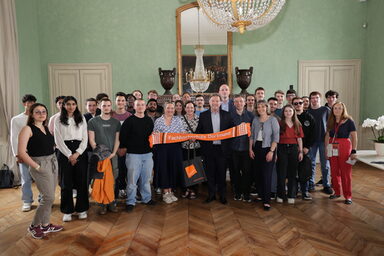 Eine Gruppe von Personen steht in einem Saal, die Personen in der Mitte halten einen orangen Schal mit der Aufschrift Fachhochschule Dortmund.