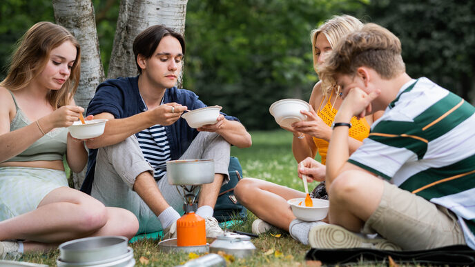 Four teenagers sit on a meadow and eat from plates.