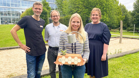 Vier Personen stehen auf dem Campus der FH Dortmund, eine Person hält einen Preis-Pokal in den Händen.