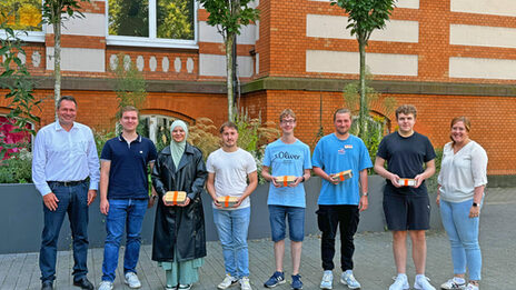 Eight people stand next to each other in front of a UAS building, some holding small boxes in front of them.