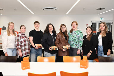 Eight people stand in a bright, large room behind a white table and look friendly into the camera.