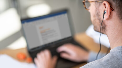 Photo of a man at laptop in home office with headphones in ear. __ Man at laptop in home office with headphones in ear.