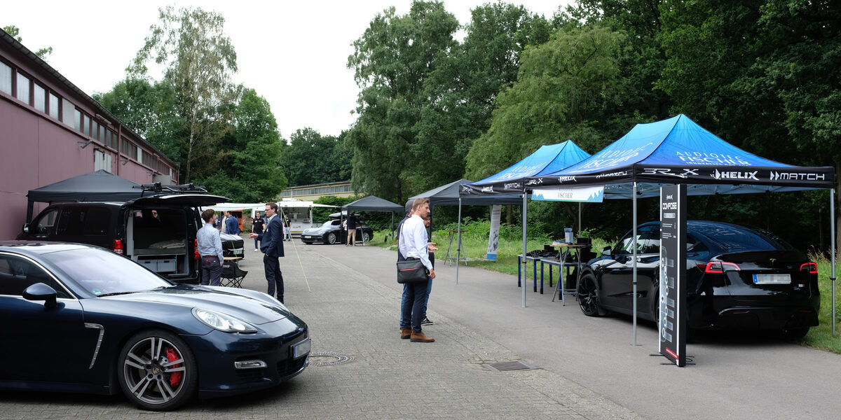 Mehrere Aussteller-Pavillons stehen an einer Straße neben einer Werkshalle, unter einigen von ihnen stehen moderne Autos, dazwischen einige Menschen.
