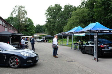 Mehrere Aussteller-Pavillons stehen an einer Straße neben einer Werkshalle, unter einigen von ihnen stehen moderne Autos, dazwischen einige Menschen.