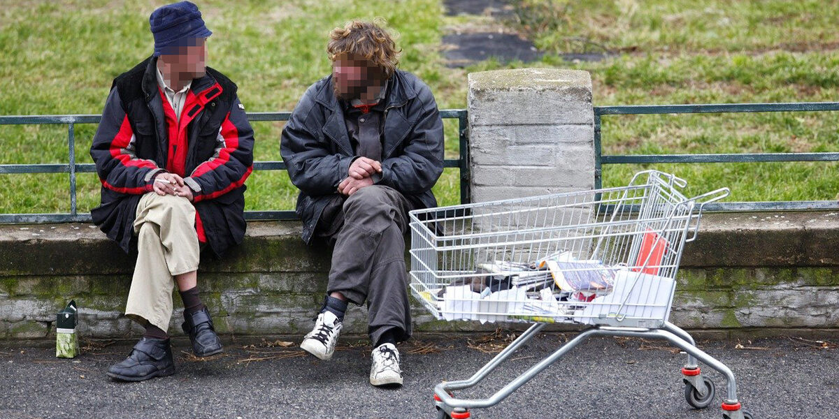 Zwei Männer sitzen auf einer Steinkante eines Parks. Vor ihnen steht ein Einkaufswagen mit ein paar Habseligkeiten.