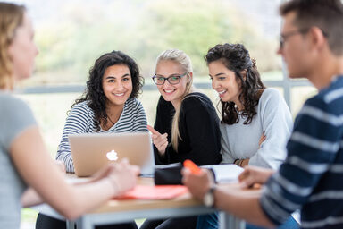 Foto von fünf Studierenden an einem Gruppentisch. Im Fokus drei Studentinnen, die nebeneinander sitzen, vor ihnen auf dem Tisch ein Laptop.